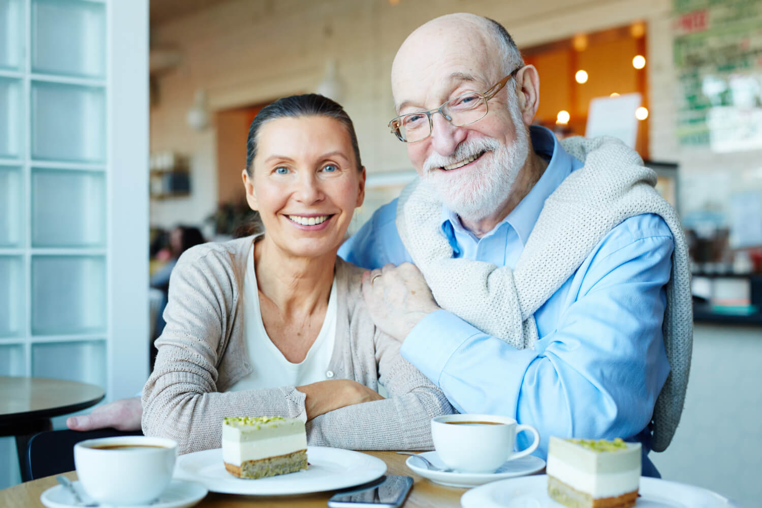 Casal idoso sorrindo em um café comendo bolo após tratamento de prótese protocolo na Clínica Arte Dental no Jd Anália Franco.
